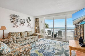 A living room with a sofa, a coffee table, a lamp, and a large window with a view of the beach.