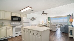 A kitchen with white cabinets, a microwave, and an oven. There is a dining area with a table and chairs, and a view of the ocean through large windows.