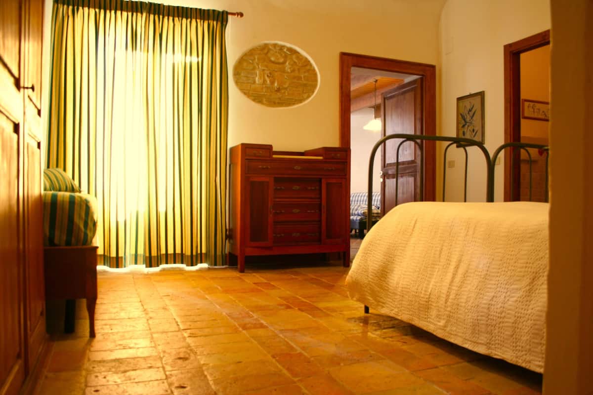 Une chambre à coucher avec un lit, une commode en bois, une table de chevet, une chaise et un rideau.