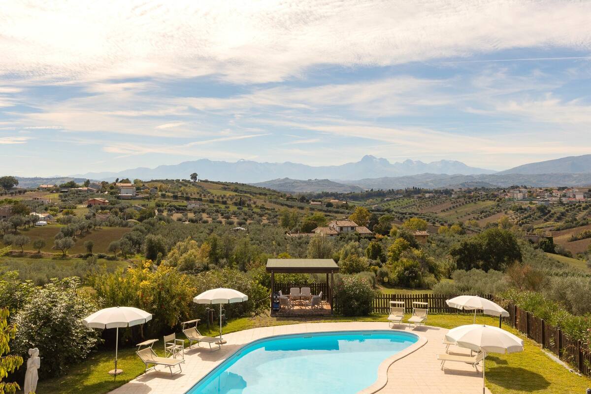 Une piscine entourée de chaises longues et de parasols, au milieu d’une végétation luxuriante, offrant une vue panoramique sur des collines et des montagnes.