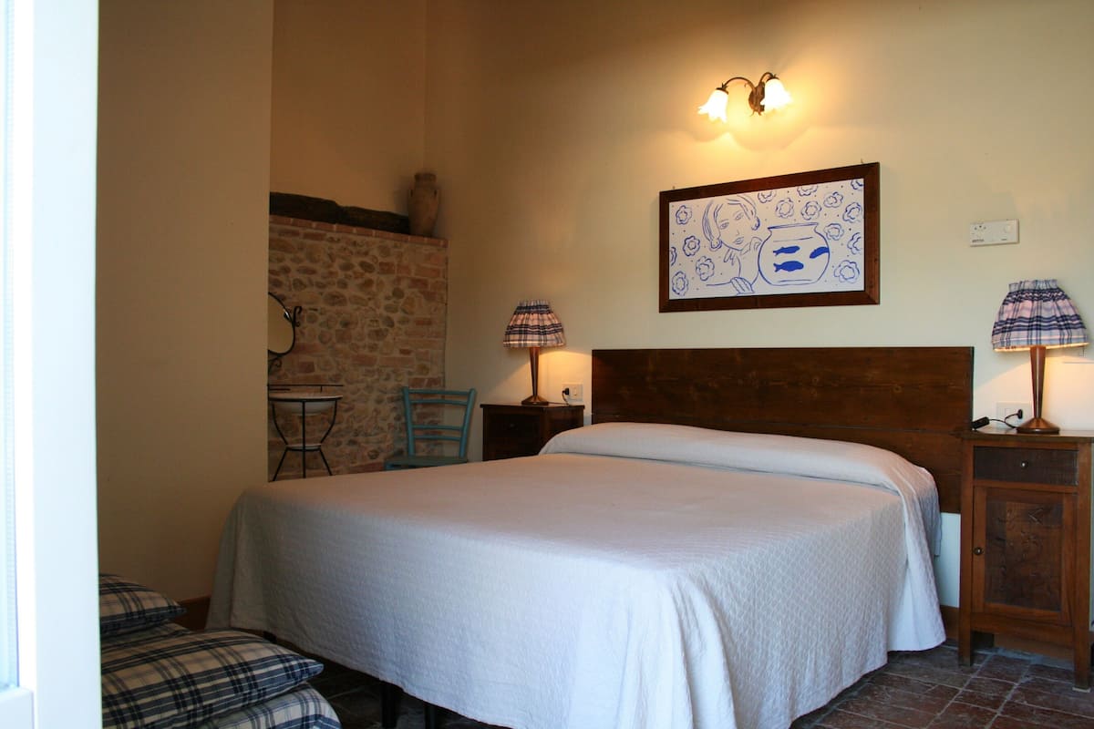 Une chambre avec un grand lit, une tête de lit en bois, un mur en pierre, une petite table, deux chaises et une table de chevet avec une lampe.