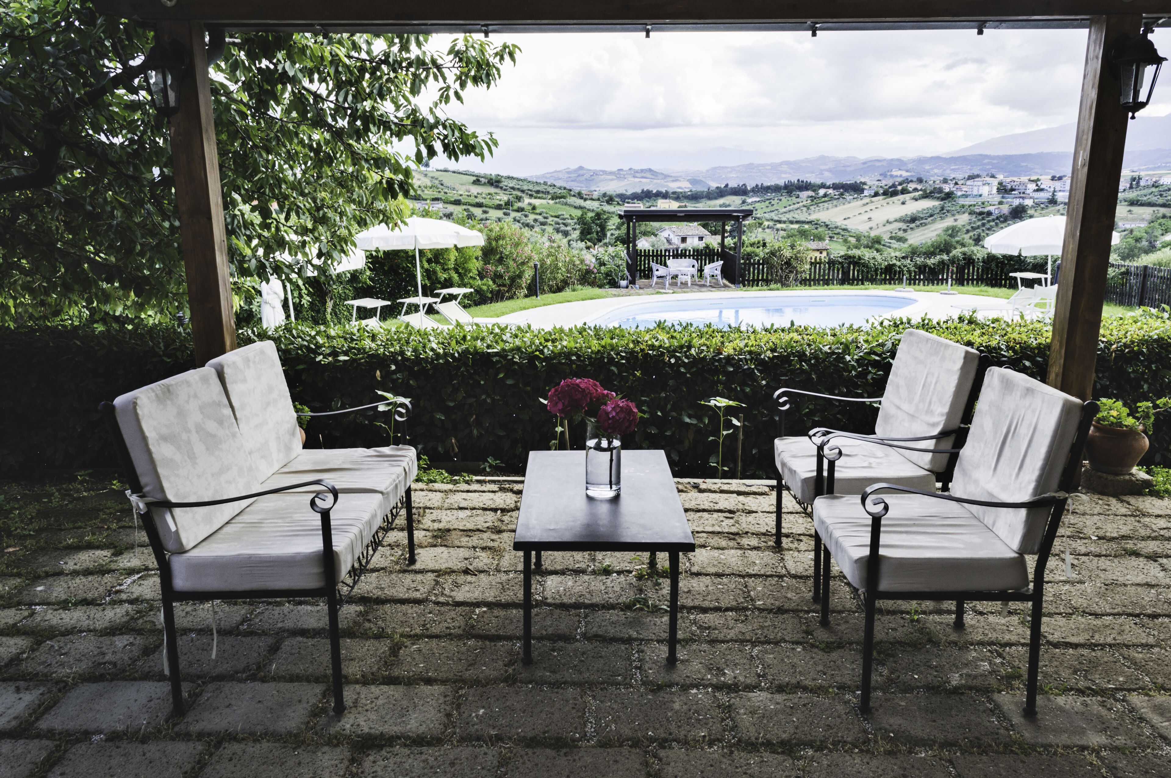 Un área de estar exterior techada con dos sillones blancos acolchados y una mesa, un florero con flores, y vistas a una piscina y el paisaje.