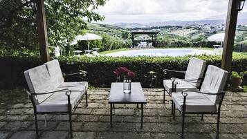 Un área de estar exterior techada con dos sillones blancos acolchados y una mesa, un florero con flores, y vistas a una piscina y el paisaje.
