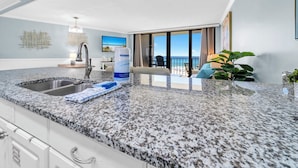 A modern kitchen with granite countertops, a stainless steel sink, and a large window offering a view of the beach.