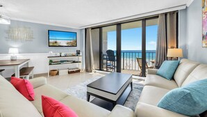 A modern living room with a sofa, coffee table, and a TV. There is a balcony with a view of the beach.