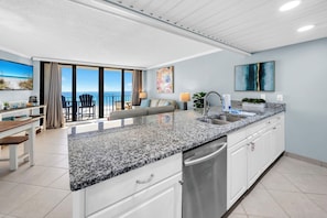 Una cocina moderna con encimeras de granito, armarios blancos y electrodomésticos de acero inoxidable. Hay una zona de comedor con mesa y sillas, y un ventanal con vistas al océano.