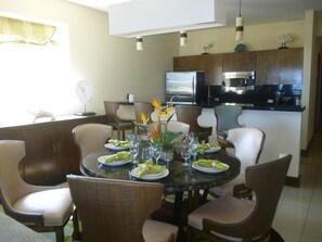 A dining area with a round glass table, surrounded by wicker chairs, and a kitchen in the background.