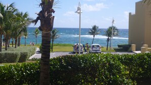 A view of a beachside area with palm trees, a road, and a building with a view of the ocean. - Spacious 1800 Sq Ft Ocean View Condo (Oyster Pond)