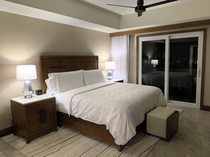 A hotel room with a large bed, two bedside tables with lamps, a wooden headboard, a sliding glass door to a balcony, and a small ottoman.