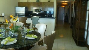 A dining area with a round table set for four, surrounded by wicker chairs. A kitchen with modern appliances is visible in the background. - Spacious 1800 Sq Ft Ocean View Condo (Oyster Pond)