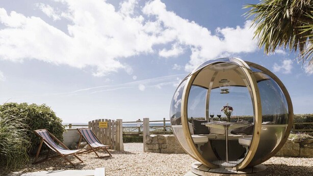 A spherical glass structure with a table and chairs inside, surrounded by a gravel area and a wooden fence. - 3 Bedroom Beachside Property with Glass Pod! (Praa Sands)