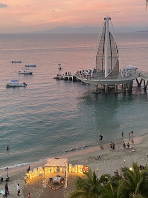 Un pont enjambant une étendue d’eau où naviguent des bateaux, une plage où se trouvent des gens, et une pancarte indiquant « JOYEUX ».
