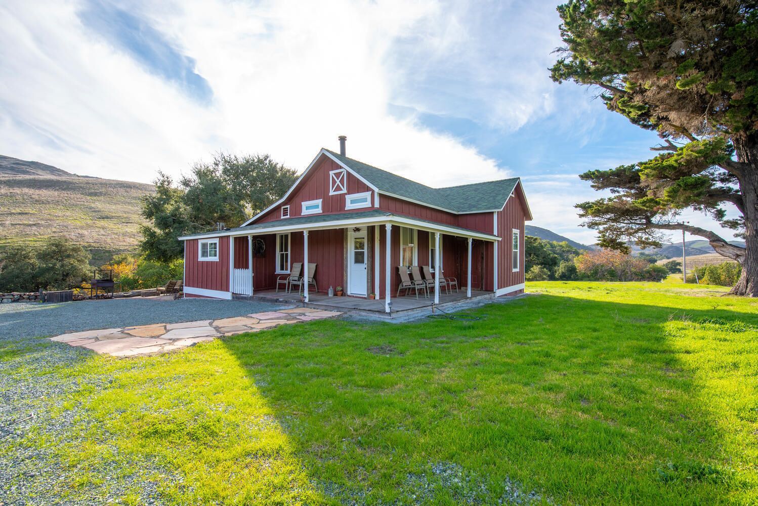Een rode, Amerikaanse boerderijstijl woning met een veranda, omringd door een gazon en bomen, met heuvels op de achtergrond.