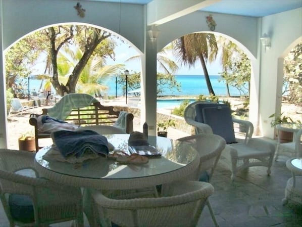 A covered outdoor seating area with wicker furniture, a glass table, and a view of the ocean through arched openings.