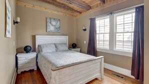 Une chambre avec un plafond en bois, un lit recouvert d’une couvre-lit à motifs, deux tables de chevet avec des lampes et une fenêtre avec des rideaux.