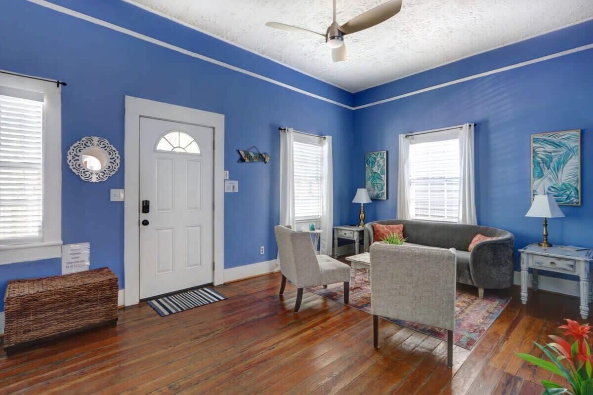 A room with blue walls, a white door, a ceiling fan, a gray sofa, a small table, and a bench.