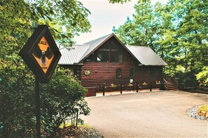A rustic log cabin with a metal roof, surrounded by trees and a gravel driveway.