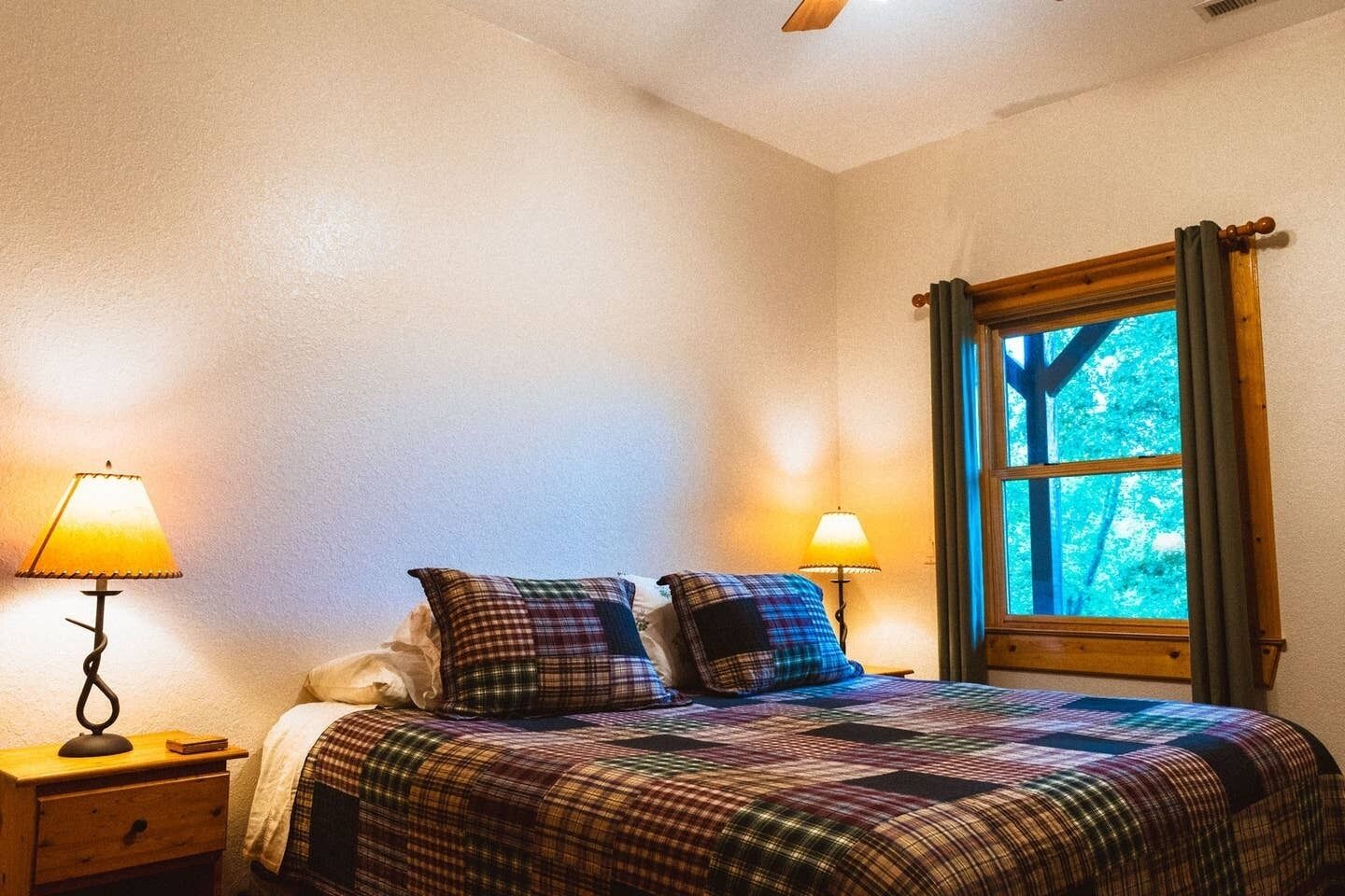 Ett sovrum med en säng, två sänglampor, ett fönster med gardiner och en sänggavel i trä.