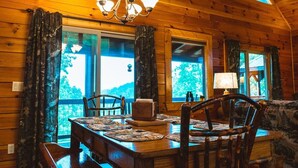 A wooden cabin interior with a dining table, chairs, a chandelier, and large windows. - Beautiful Views of The Great Smoky Mountains From Anywhere Indoors or Out! (Bryson City)