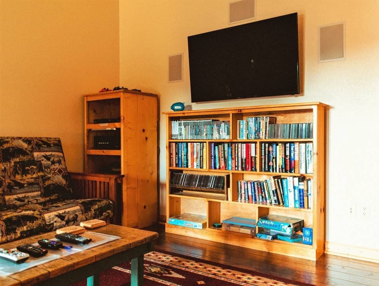 A living room with a couch, a coffee table with remotes, a bookshelf, and a television on the wall.