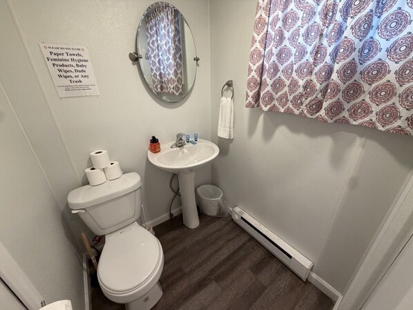 A bathroom with a toilet, sink, and mirror. There is a sign on the wall and a patterned shower curtain.