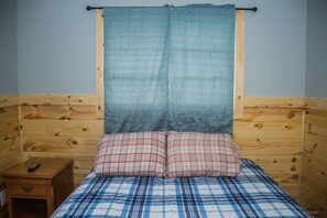 Ein Einzelbett mit karierter Tagesdecke, zwei karierte Kissen, ein Holzkopfstück und ein kleiner Nachttisch mit Fernbedienung.