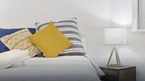 Un lit bien fait, avec une literie à rayures et un oreiller jaune, une table de chevet avec une lampe et une fenêtre avec des stores.