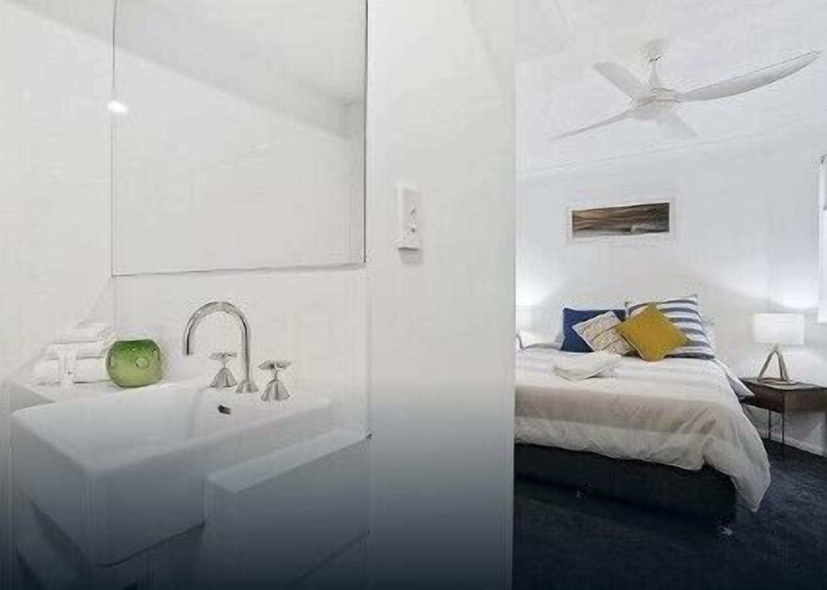 A modern bathroom with a large mirror, a sink, and a green soap dispenser. The room has a white color scheme and a ceiling fan.
