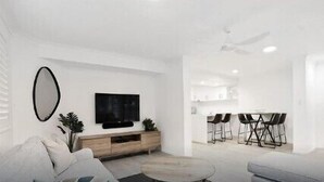 Un salon moderne avec un canapé, une table basse et un téléviseur fixé au mur. On aperçoit à l’arrière-plan un coin repas avec une table et des chaises.