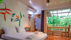 A hotel room with a bed, a wooden bench, a window with a view of greenery, a bathroom with a shower curtain, and an air conditioning unit.