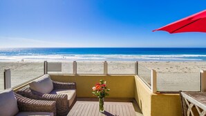 A balcony with wicker furniture and a vase of flowers, overlooking a beach with a clear blue sky.