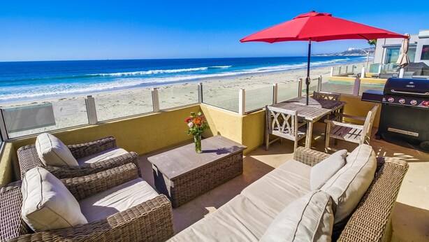 A balcony with wicker furniture, a red umbrella, and a view of the beach and ocean.