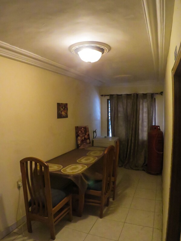 Un coin repas avec une table dressée pour quatre personnes, des chaises en bois, un luminaire au plafond, un tableau accroché au mur et un objet rouge dans un coin.