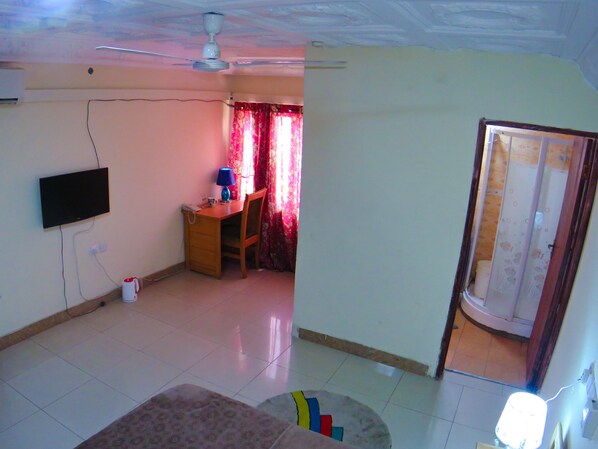 A room with a ceiling fan, a flat-screen TV mounted on the wall, a desk with a chair, and a bathroom visible through an open door. - St Mary's at hills guest house (Janman)