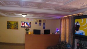 Zona de recepción de hotel con techo de molduras decorativas, un mostrador de recepción con una computadora portátil, un televisor y un reloj de pared.