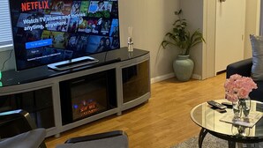 Un salón con una televisión de pantalla plana sobre un mueble bajo, una mesa de centro de cristal con un jarrón de flores, un sofá gris y suelo de madera.