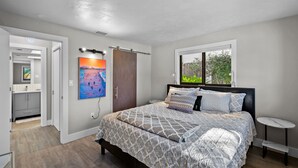 Een slaapkamer met een groot bed, een raam, een nachtkastje en een deur naar een andere kamer.