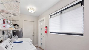 Een wasserette met een wasmachine, planken met schoonmaakmiddelen, een brandblusser en een raam met jaloezieën.