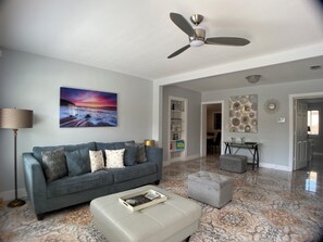 A living room with a grey sofa, a white ottoman, a ceiling fan, a painting of a sunset, and a tiled floor.