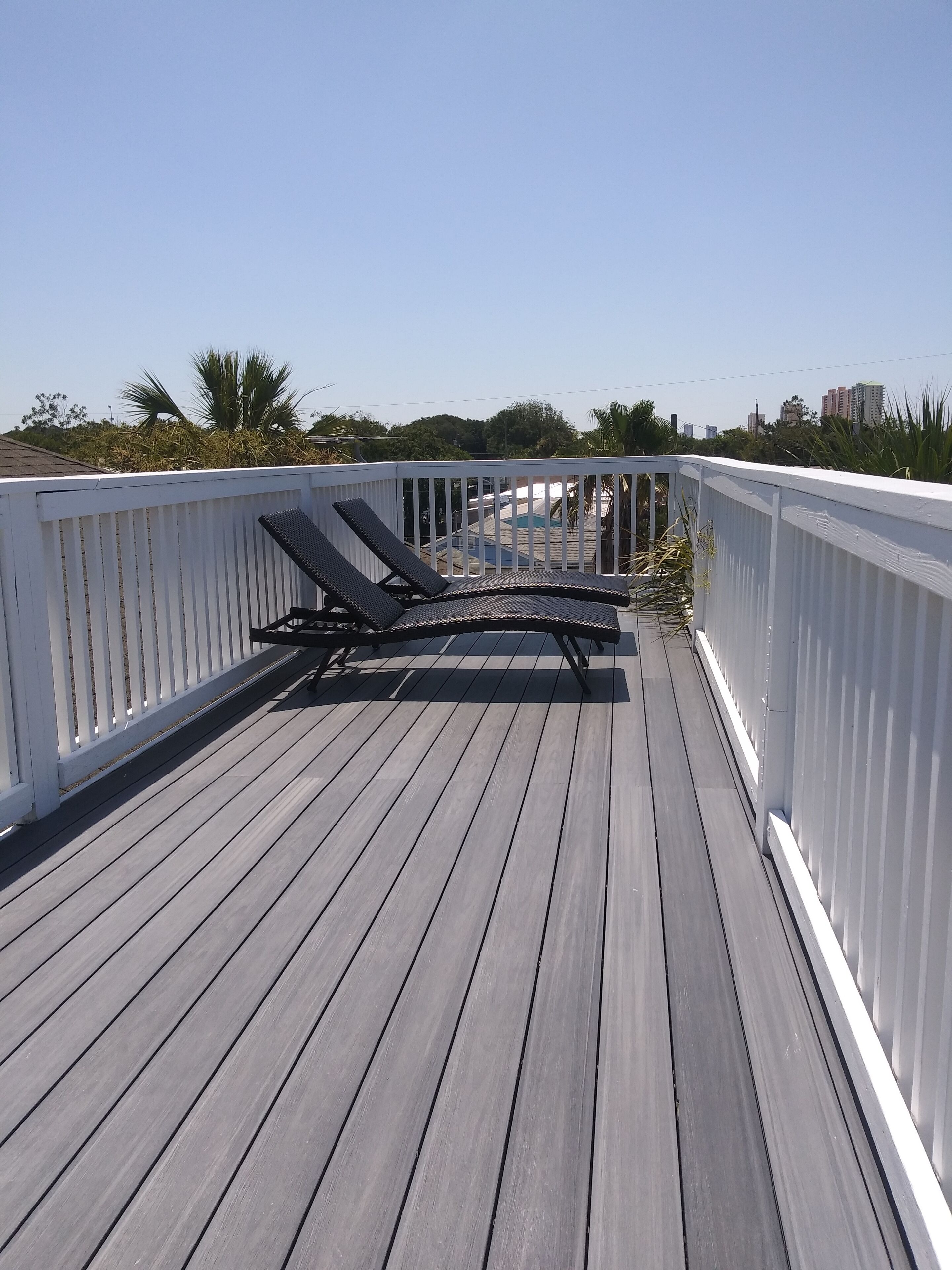 A wooden deck with two lounge chairs, white railings, and a view of a swimming pool and buildings in the background.