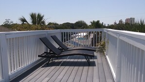 Eine Holzterrasse mit zwei Liegestühlen, weißen Geländern und Blick auf einen Swimmingpool sowie Gebäude im Hintergrund.