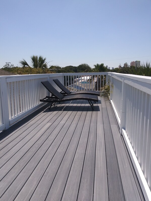 A wooden deck with two lounge chairs, white railings, and a view of a swimming pool and buildings in the background.
