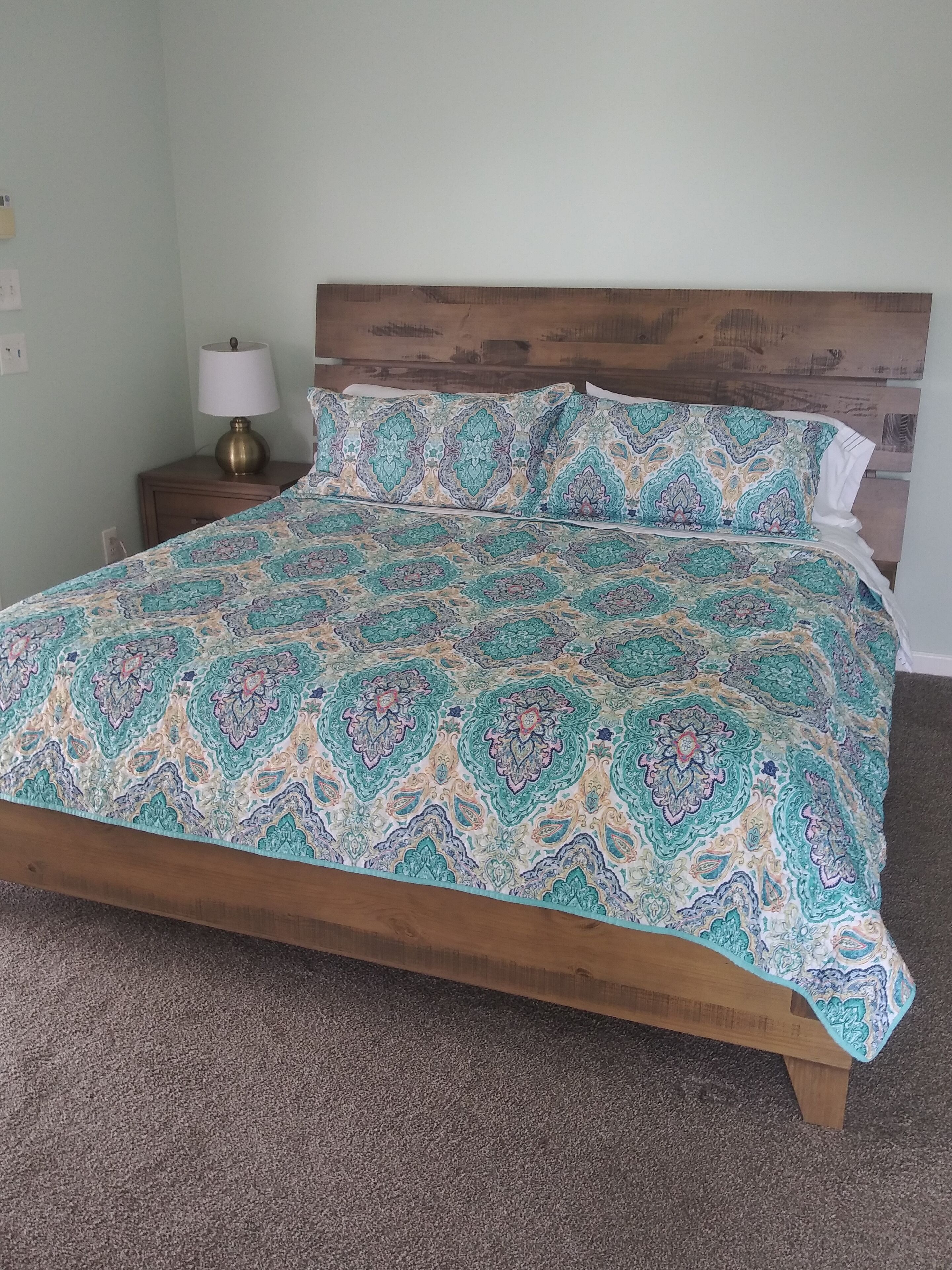 Ein Bett mit gemustertem Überwurf und Kissen, ein Holz-Kopfschild, ein Nachttisch mit Lampe und ein Teppichboden.