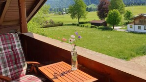 A wooden balcony with a plaid cushion and a wooden table, overlooking a green landscape with trees and a mountain in the background. - Apartment Panorama (Reit im Winkl)