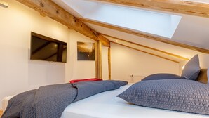 A bedroom with a sloped ceiling, wooden beams, a skylight, a bed with grey bedding, and a mounted TV. - Apartment Panorama (Reit im Winkl)
