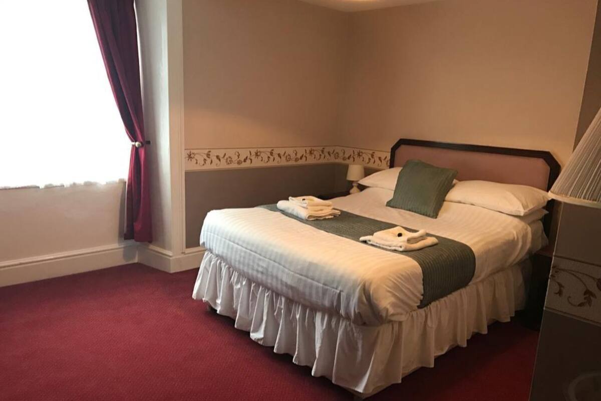 A neatly made bed with white bedding and a green throw, a bedside lamp, and a red carpeted floor.