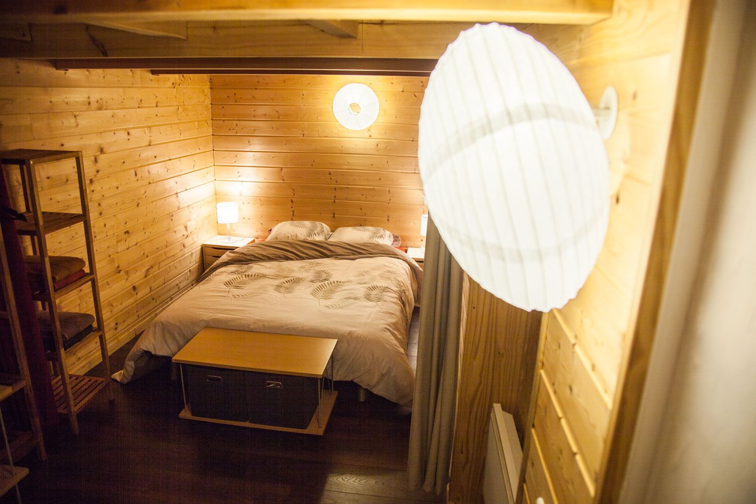 A cozy wooden room with a bed, a nightstand, a lamp, and a shelf.