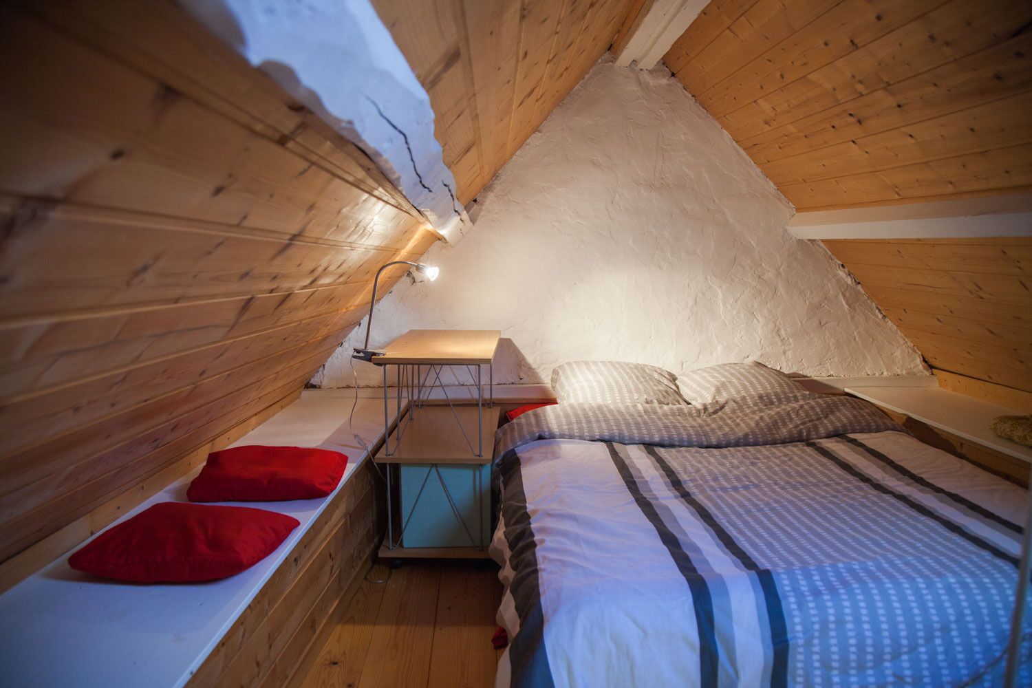A cozy attic bedroom with a sloped ceiling, a bed with a striped duvet, a nightstand with a lamp, and a small table.