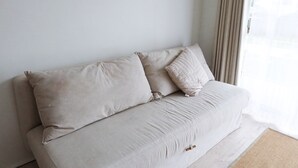 A beige sofa with two cushions against a plain wall, a window with sheer curtains, and a light-colored carpet. - The Heart of Huskisson (Huskisson)