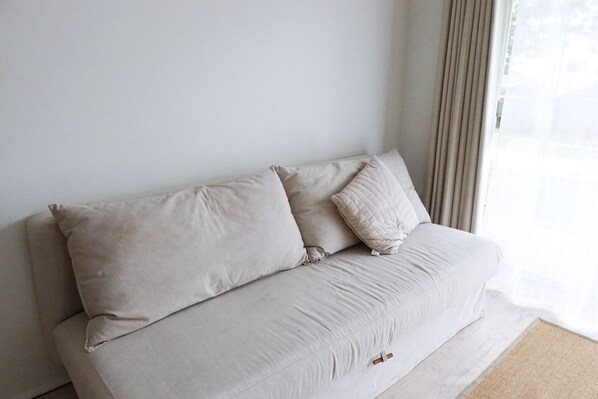 A beige sofa with two cushions against a plain wall, a window with sheer curtains, and a light-colored carpet.